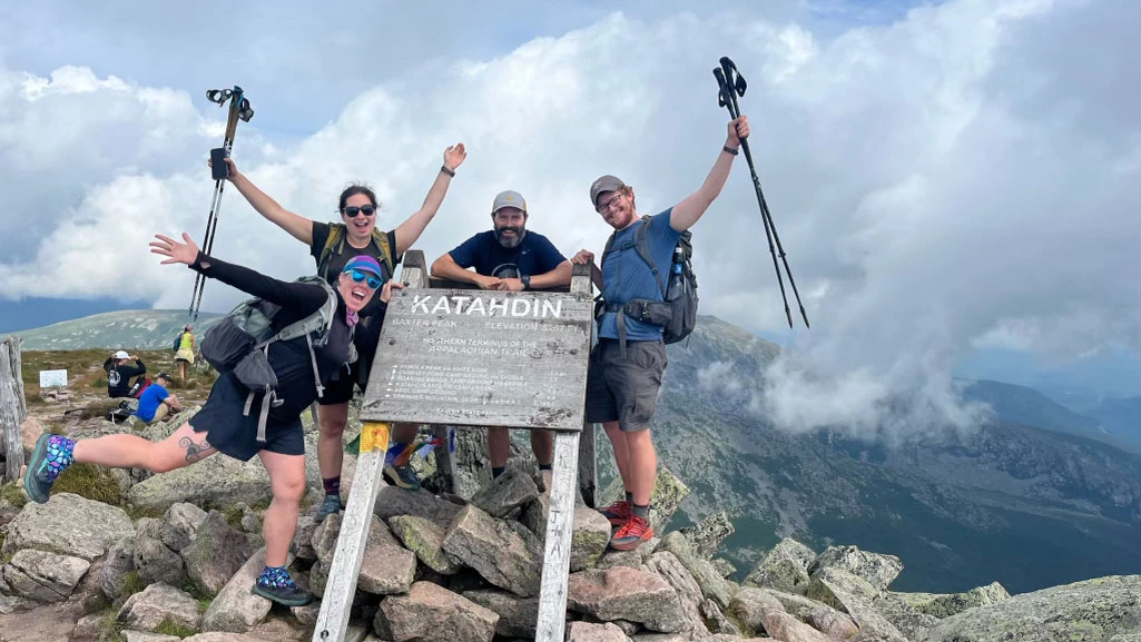 Four hikers celebrating beside the Katahdin summit sign, with mountains and clouds behind them