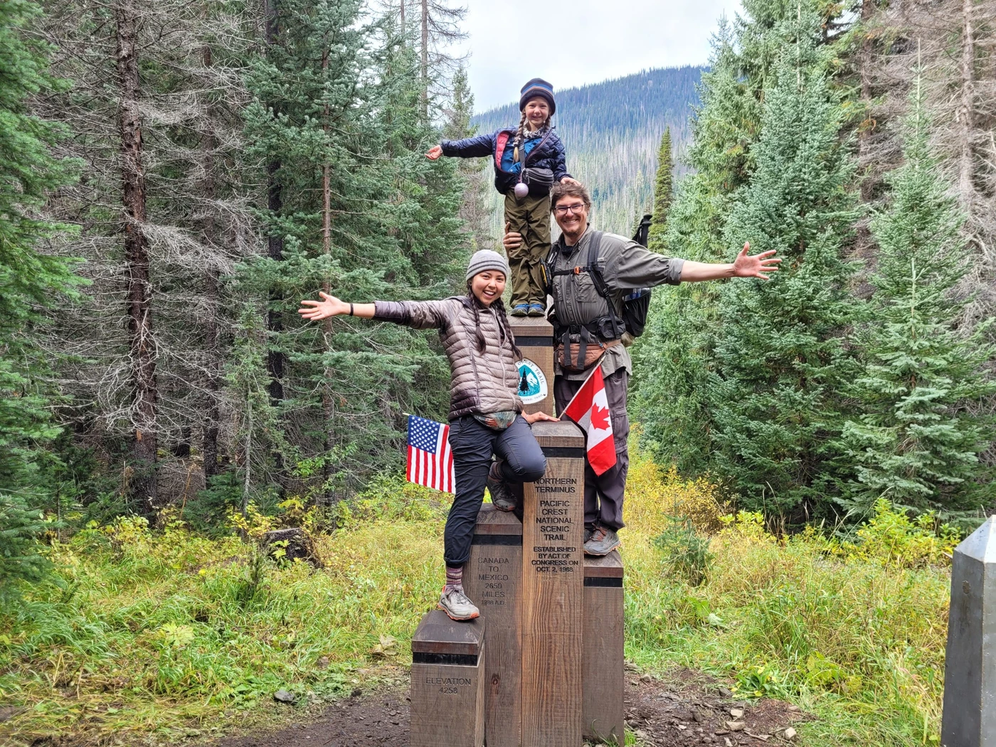 Three hikers celebrating at the Pacific Crest Trail northern terminus monument in the forest